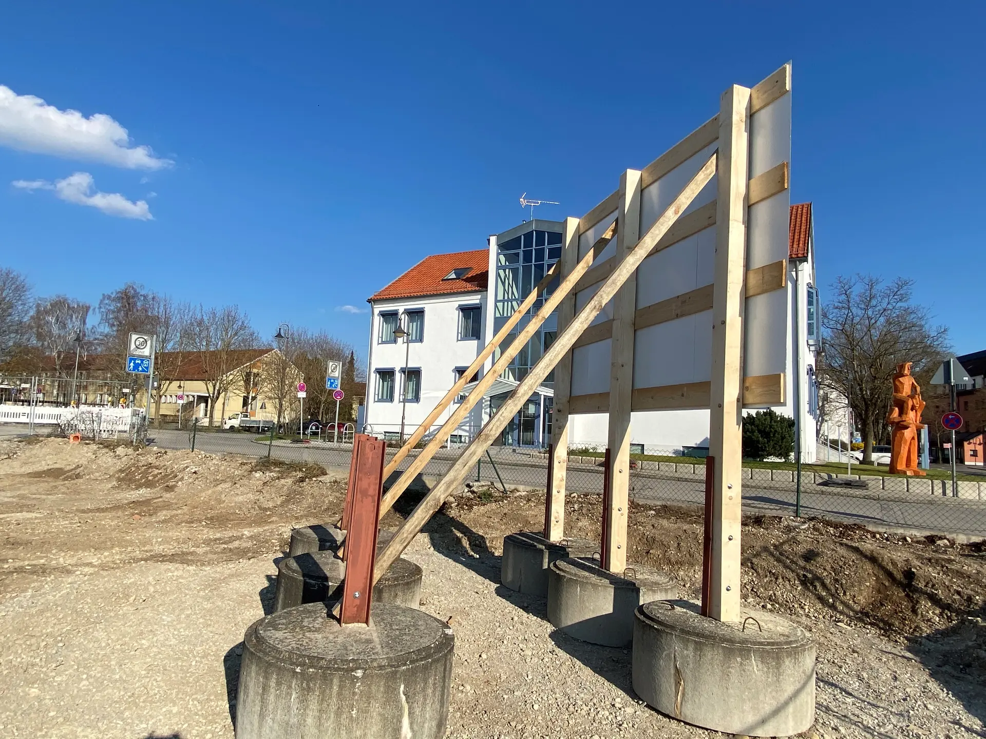 Werbetechnik Dümpel - Augsburg | Großes Außenschild-Holzbalken-Betonsockel-Kiesfläche-Straßenansicht-Gebäude-Skulpturen-Stadtlandschaft.