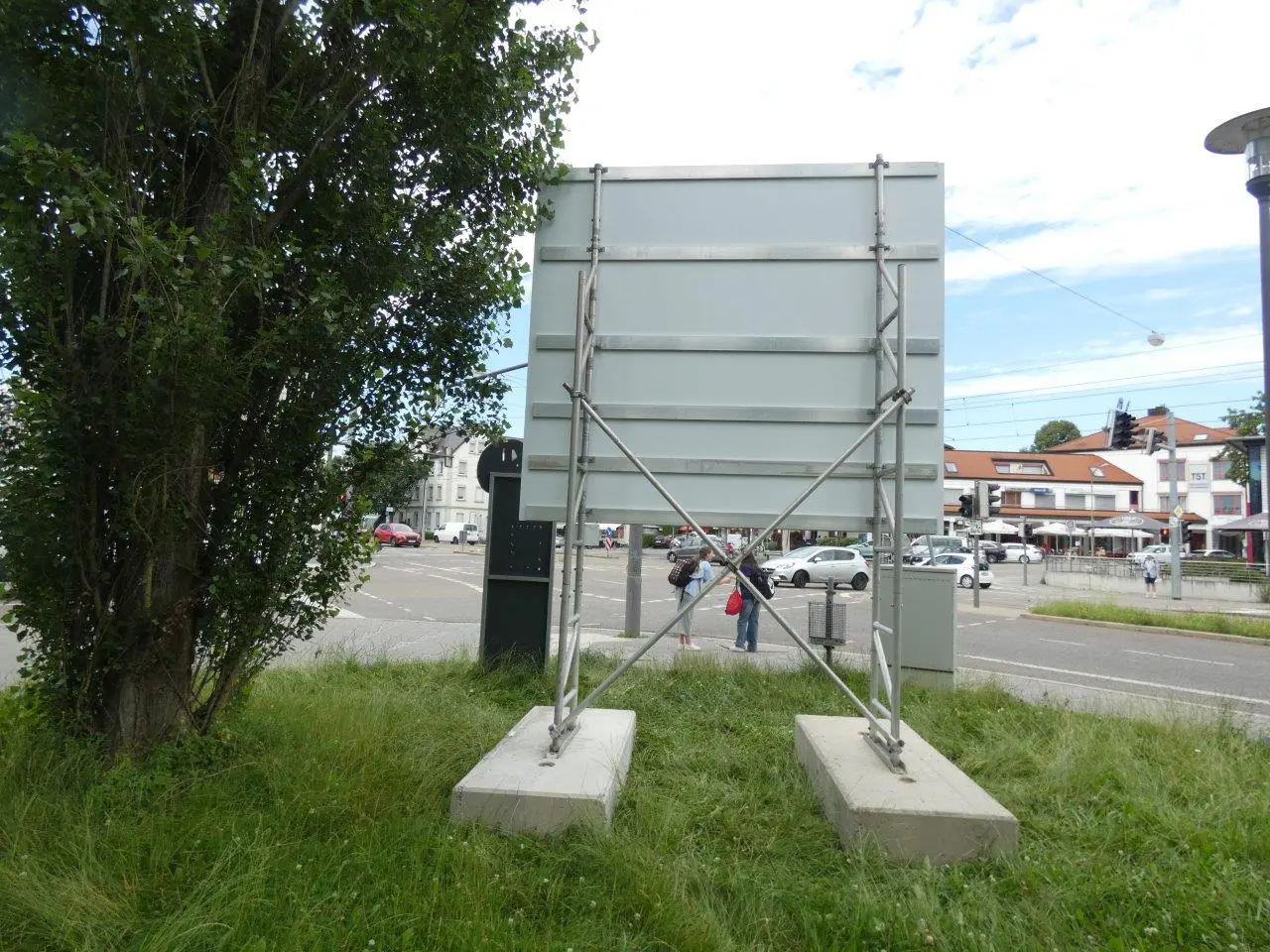 Werbetechnik Dümpel - Augsburg | Blank Plakatwand auf Betonsockel - Stadt Straße - grasbewachsenen Bereich - städtischen Werbung Bord - Gebäude - Fußgänger im Hintergrund.