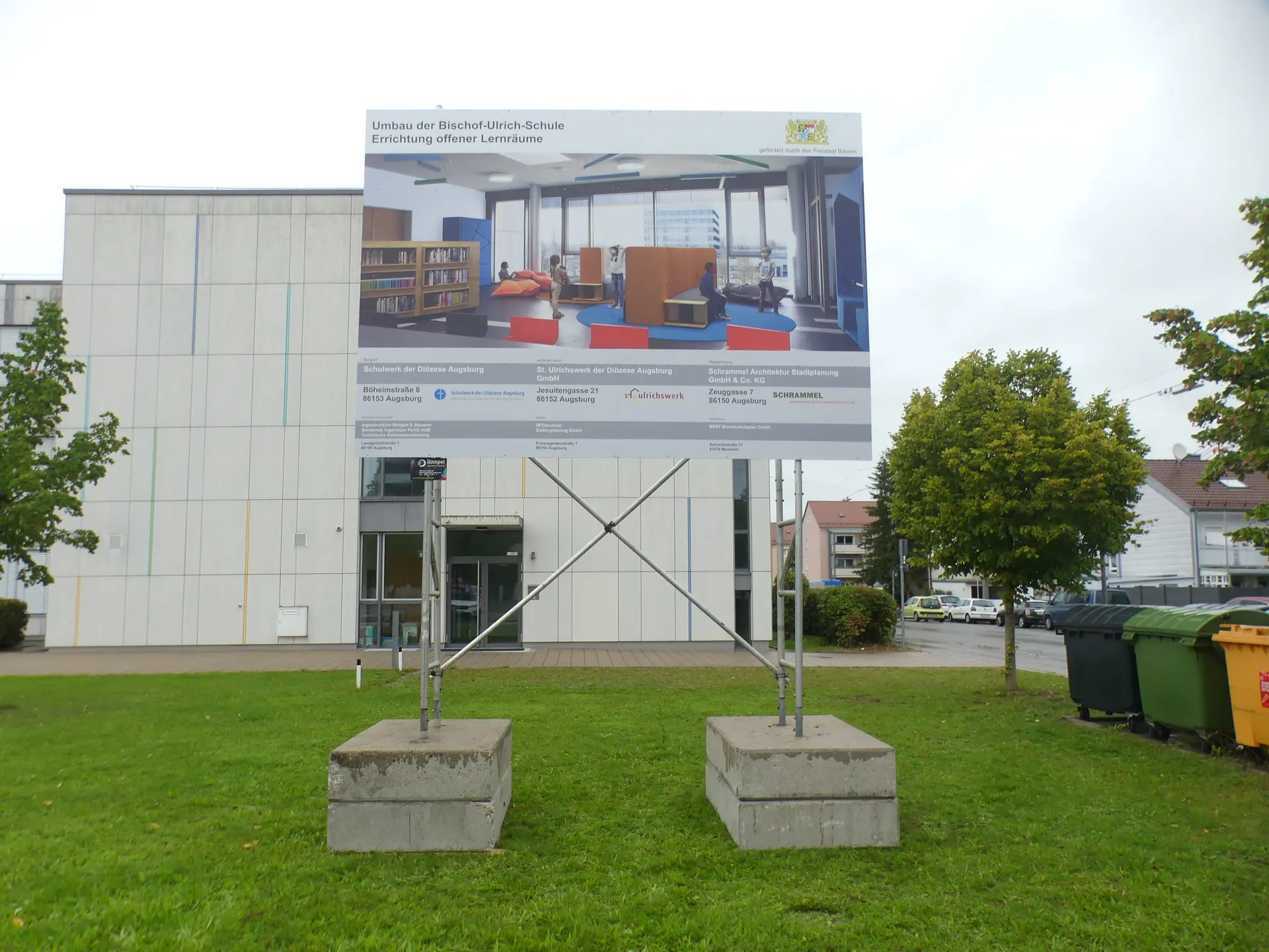 Werbetechnik Dümpel - Augsburg | Schild mit architektonischen Plänen - modernes Äußeres des Gebäudes - Landschaftsgestaltung mit Bäumen und Rasen - sichtbare Recycling-Behälter.