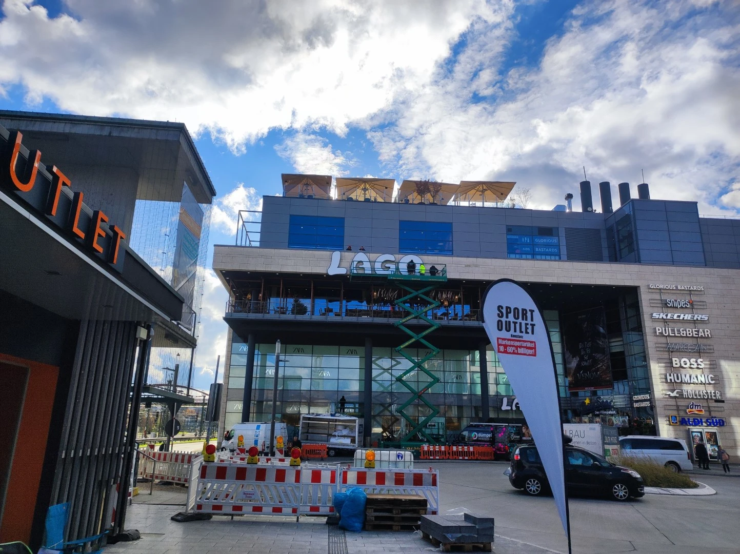 Werbetechnik Dümpel - Augsburg | Modernes Einkaufszentrum - Glasfassade - überdachte Freisitze - Markenschilder - Bauaufzug - teilweise bewölkter Himmel - städtisches Einkaufszentrum.