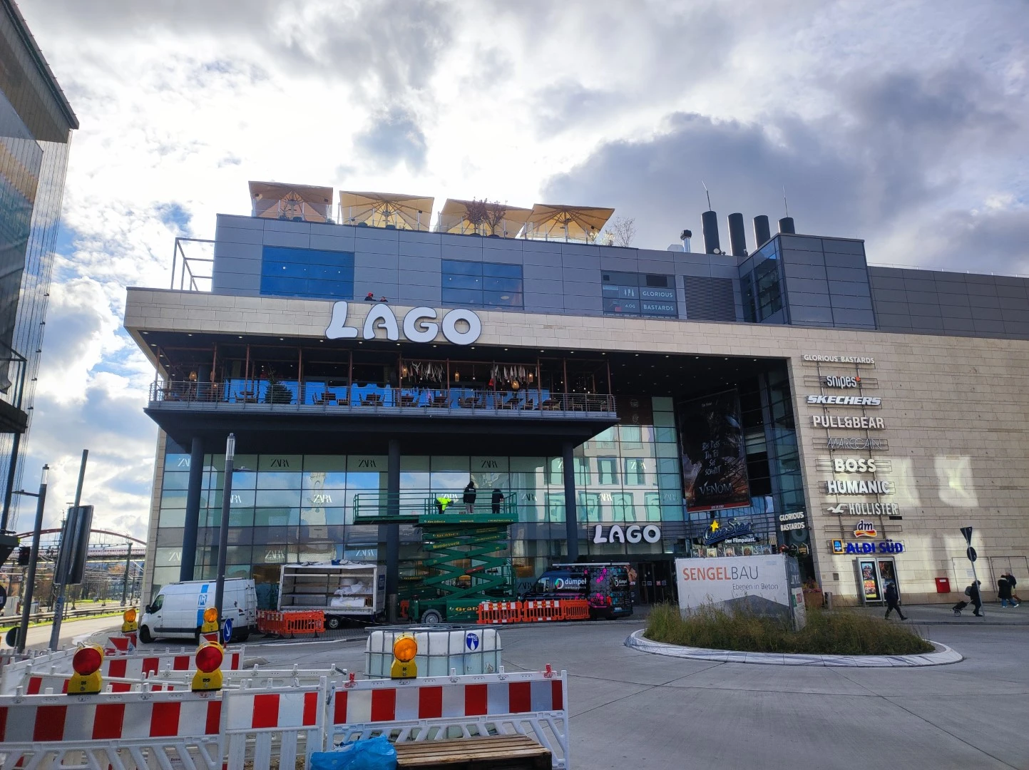Werbetechnik Dümpel - Augsburg | LAGO Einkaufszentrum - Glasfassade - Sitzgelegenheiten im Freien - Einzelhandelsgeschäfte - Bauabsperrungen - bewölkter Himmel.