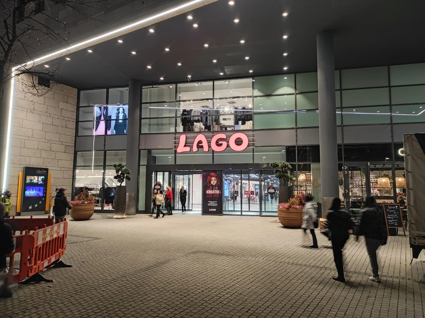 Werbetechnik Dümpel - Augsburg | Moderner Gebäudeeingang bei Nacht - große Glasfenster - rosafarbenes LAGO-Schild - vorbeilaufende Fußgänger - städtische Ausgehszene.