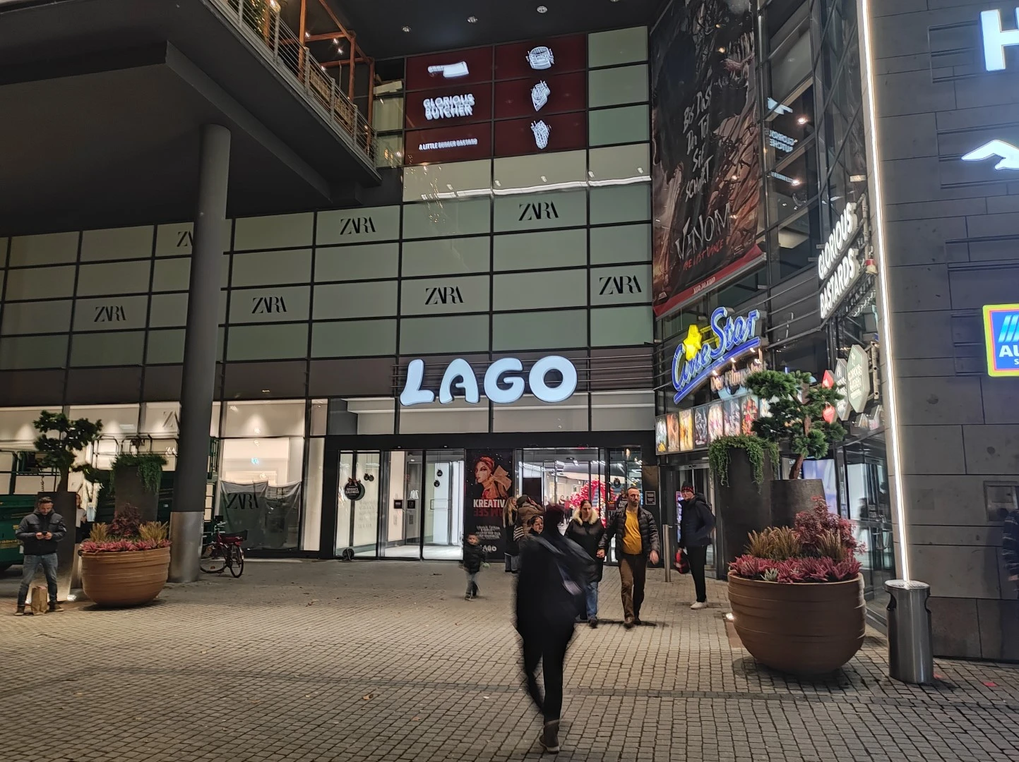 Werbetechnik Dümpel - Augsburg | LAGO-Einkaufszentrum bei Nacht - Menschen, die spazieren gehen - beleuchtete ZARA-, Casino Sport- und Aldi-Schilder - große Pflanzkübel - Schaufensterfronten.