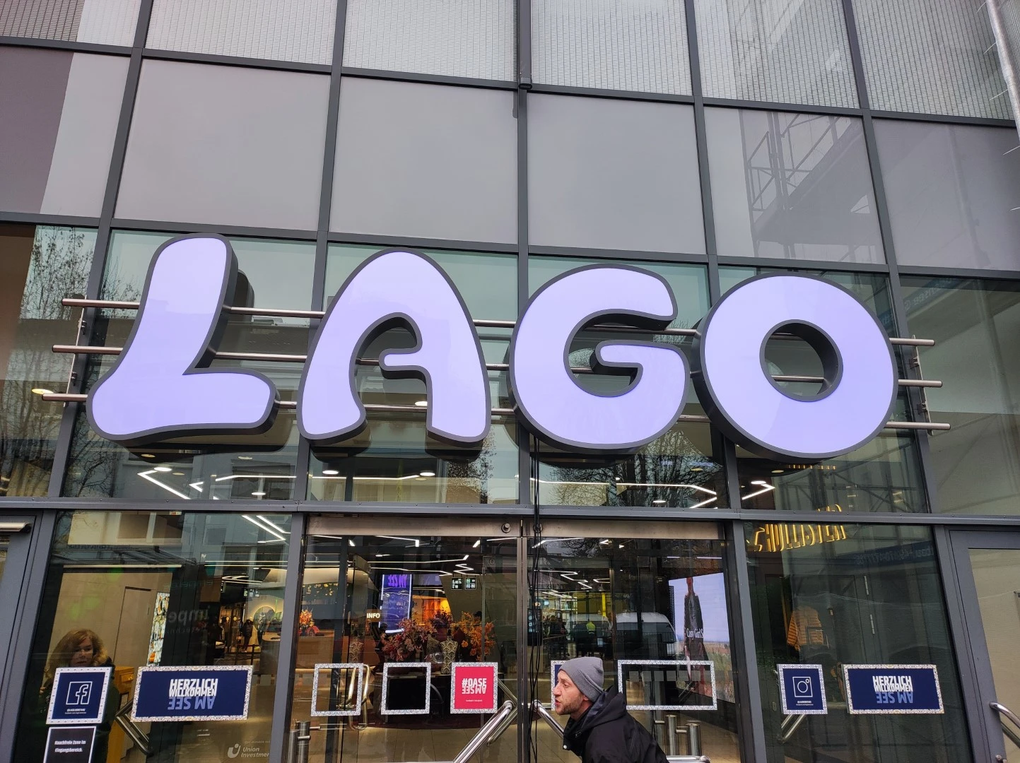 Werbetechnik Dümpel - Augsburg | LAGO-Schild über dem Eingang eines modernen Glasgebäudes - Person mit grauer Mütze im Vorbeigehen - Schaufensterbeschriftung - städtische Architektur.