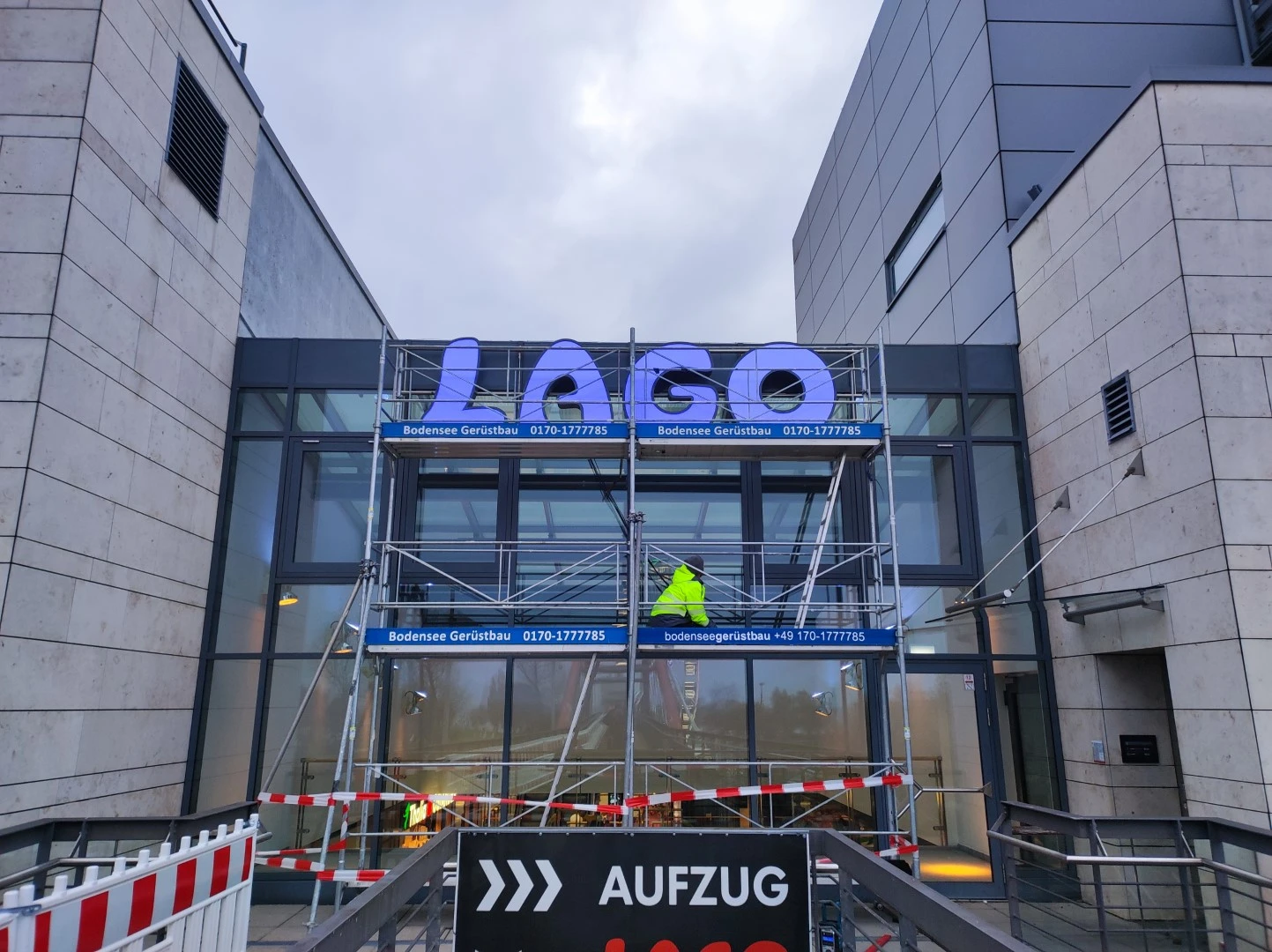 Werbetechnik Dümpel - Augsburg | Arbeiter montiert LAGO-Schild am Gebäudeeingang - Gerüst - gelbe Jacke - violette Buchstaben - Baustelle - Glastüren - Sicherheitsband.