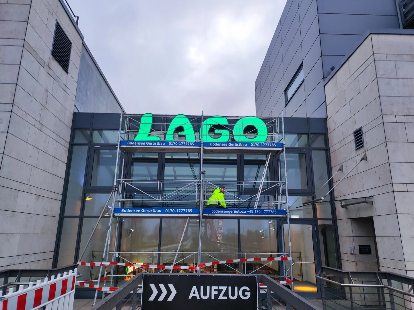 Werbetechnik Dümpel - Augsburg | LAGO Eingangsgerüst - Bauarbeiter in gelber Jacke - Wartungsarbeiten - Aufzugsschild - Gebäudesanierung.