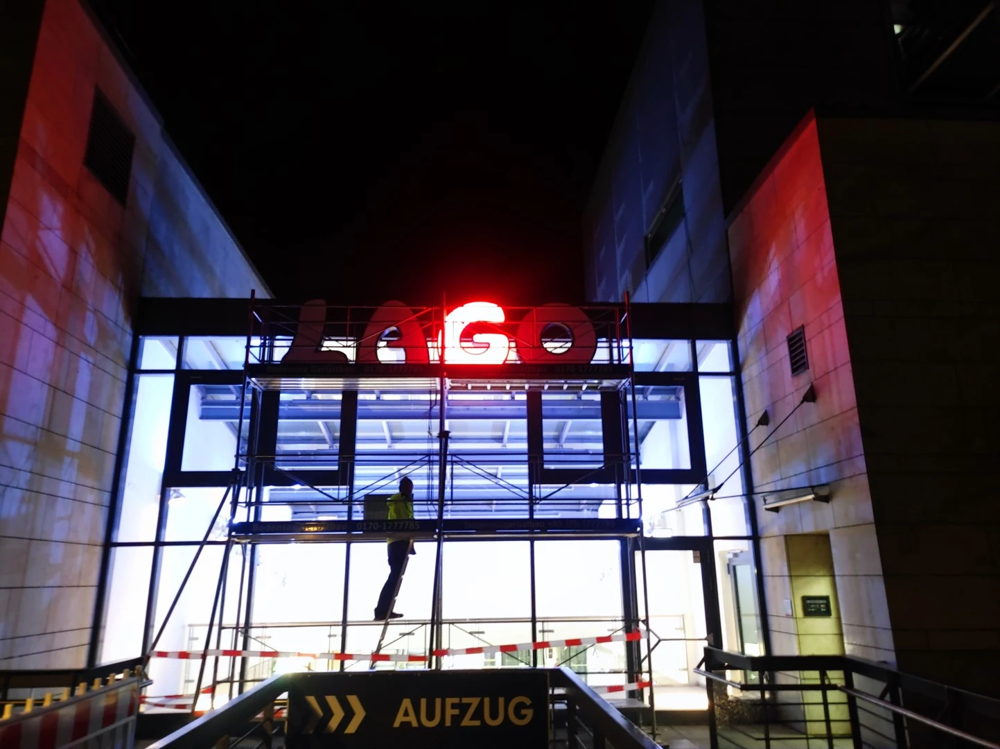 Werbetechnik Dümpel - Augsburg | Arbeiter auf einem Gerüst bei Nacht unter dem beleuchteten LAGO-Schild - Glaseingang - AUFZUG-Aufzugsschild - Einkaufszentrum außen.