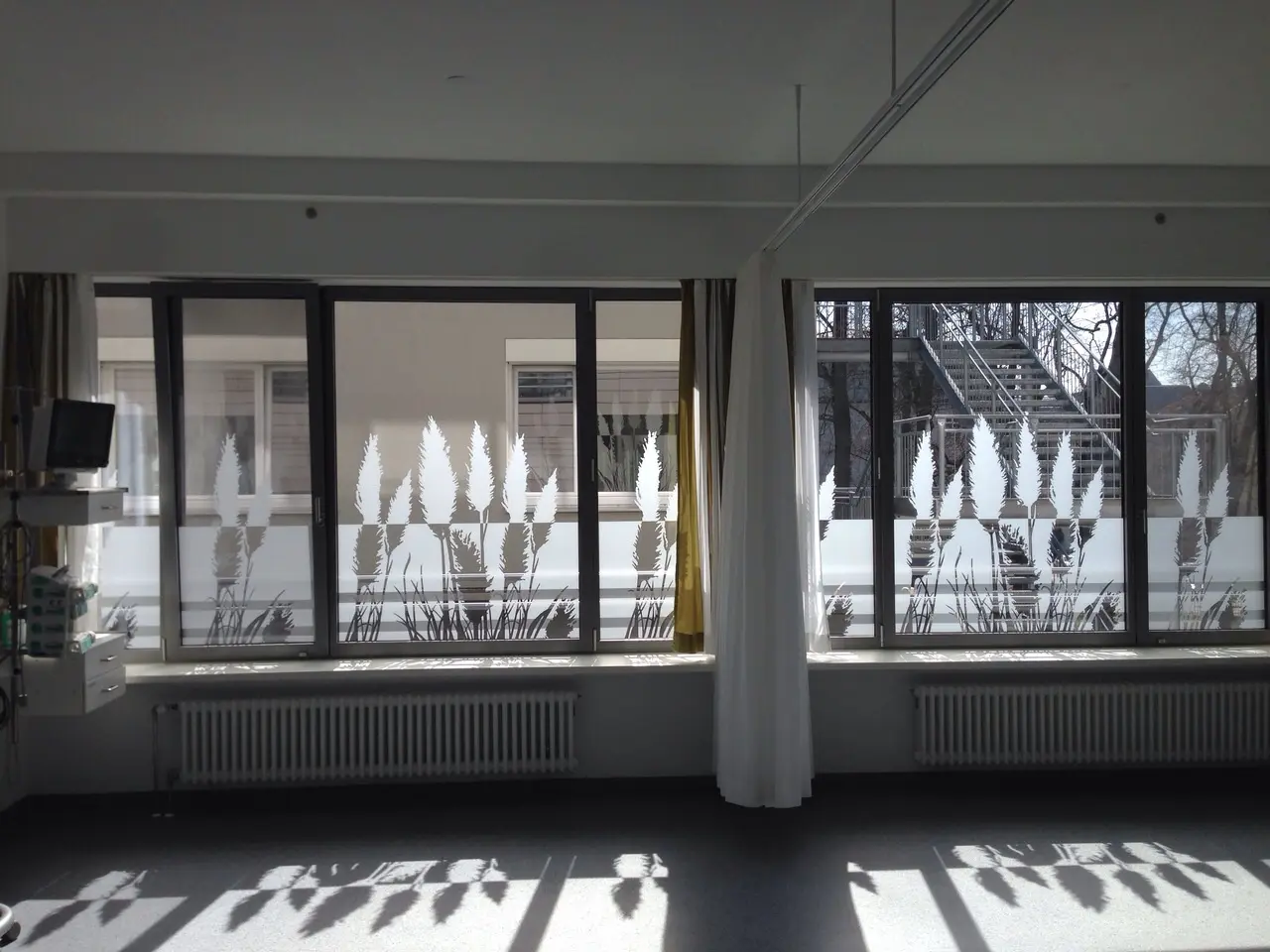 Werbetechnik Dümpel - Augsburg | Modernes Krankenhauszimmer-große Fenster-durchsichtige Vorhänge-aufgetaute Pflanzenmuster-natürliches Sonnenlicht-Schatten auf dem Boden-Gesundheitseinrichtung.