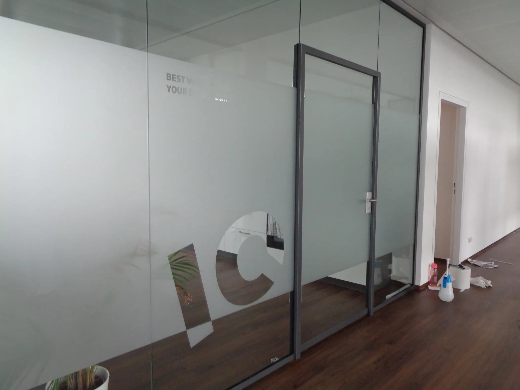 Werbetechnik Dümpel - Augsburg | Gläserne Bürotür-IC-Buchstaben-teilweise weißes, gefrostetes Design-Reinigungsmittel auf dem Boden-Pflanze durch Glas sichtbar-moderner Arbeitsbereich.
