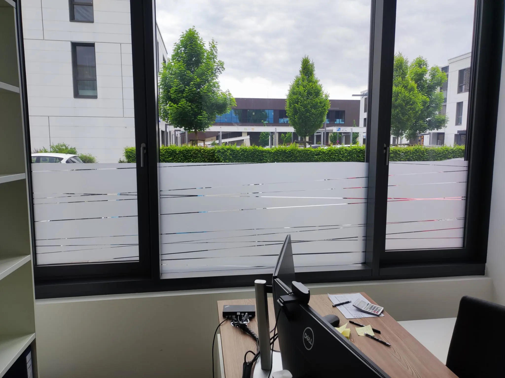 Werbetechnik Dümpel - Augsburg | Moderner Büroarbeitsplatz - Schreibtisch - Computer - großes Fenster - matte Streifen - Blick auf ein städtisches Gebäude - Grünflächen draußen.
