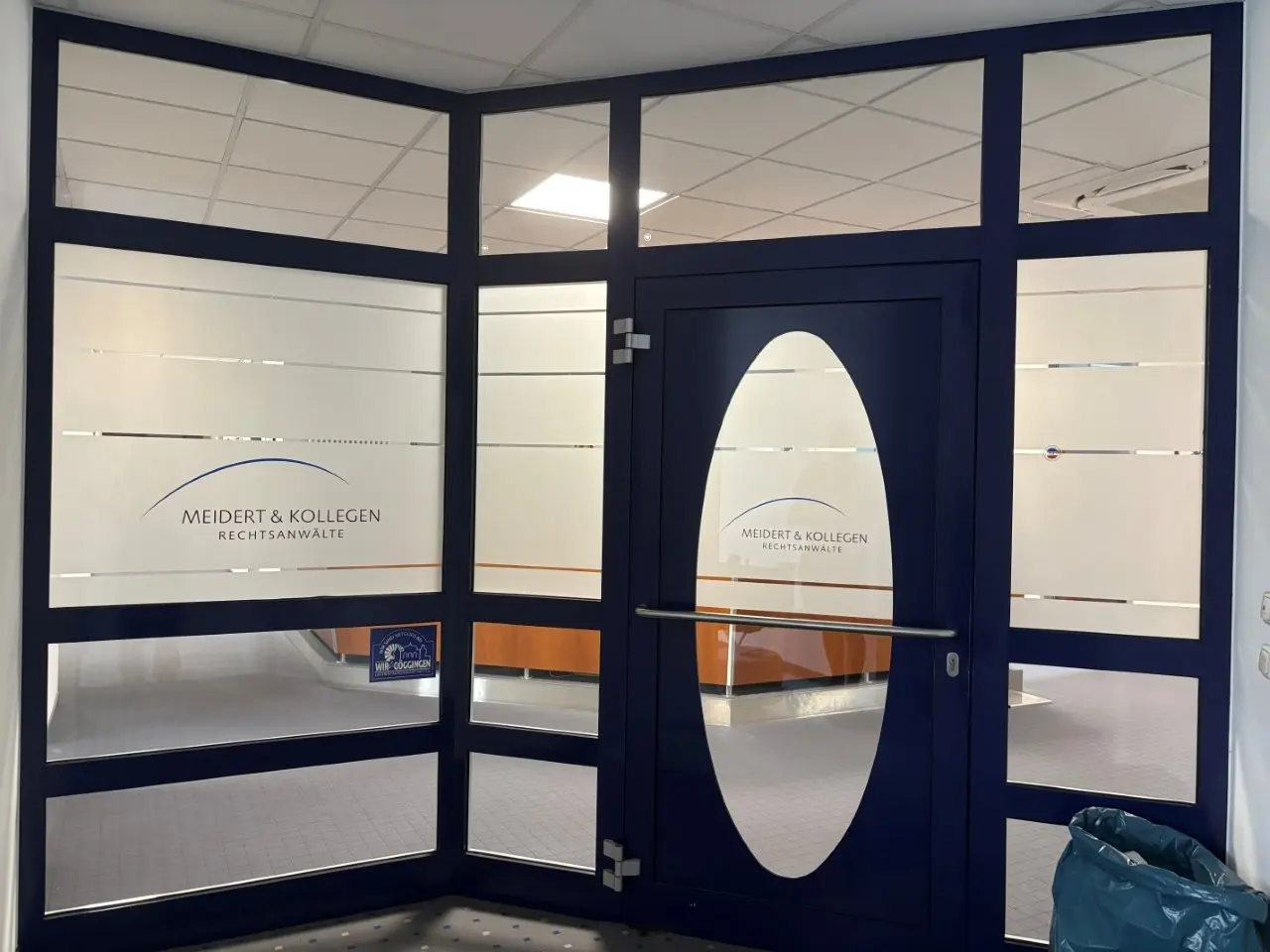 Werbetechnik Dümpel - Augsburg | Gläserner Büroeingang - Meidert & Kollegen Rechtsanwälte - Kanzlei - blauer Rahmen - mattierte Paneele - Holztresen - moderner Arbeitsbereich.